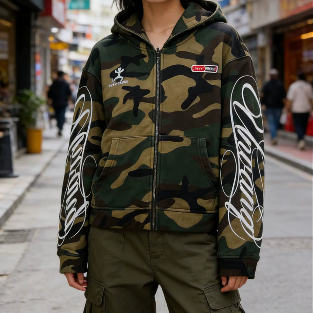 A person wearing a zip-up hoodie with a green and brown camouflage pattern, featuring large white script graphics on both sleeves and a small red-and-white chest patch, photographed on a city street.