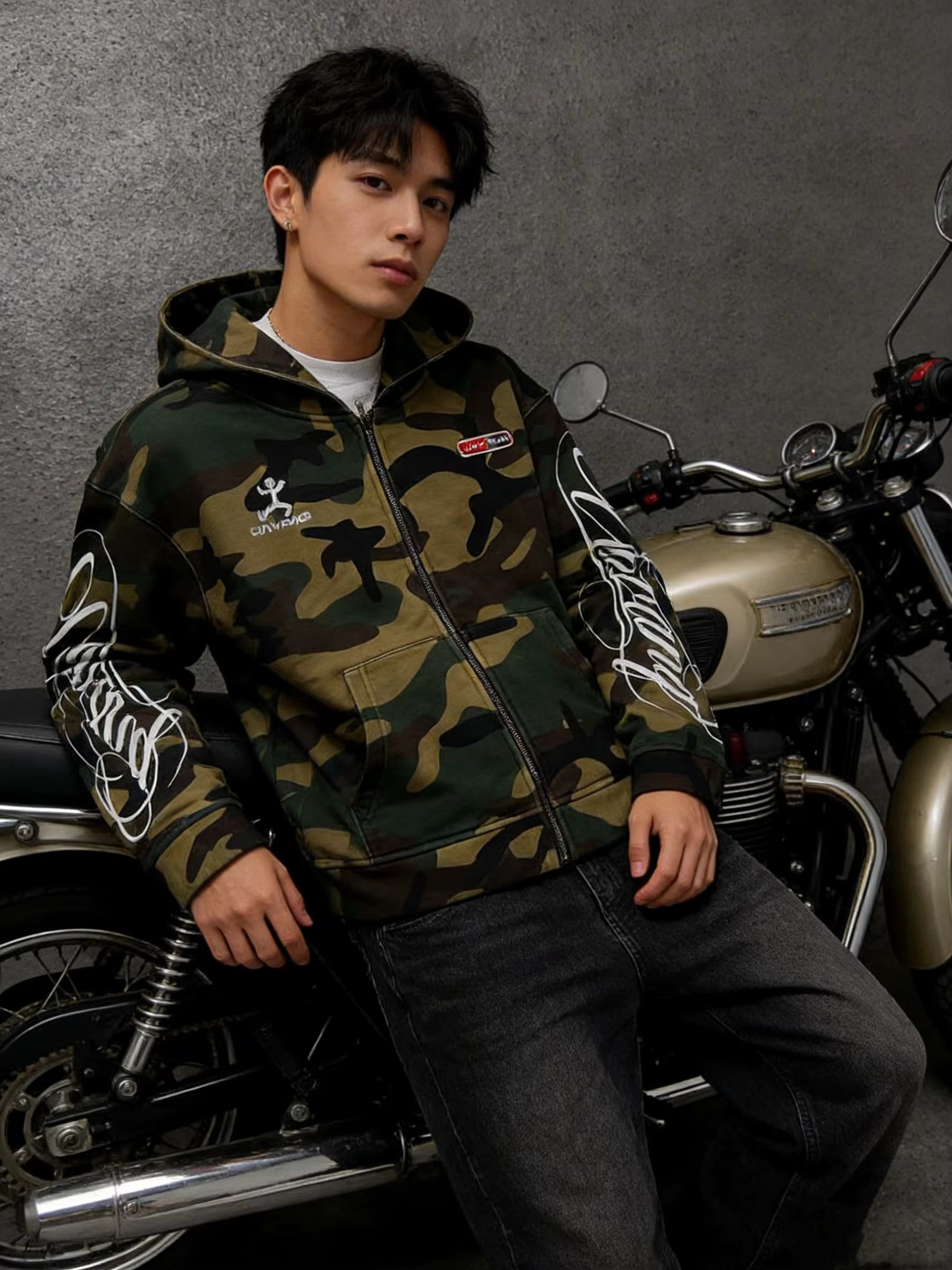 A man wearing a green camouflage zip-up hoodie with white graphic prints on the sleeves, sitting on a vintage motorcycle against a textured gray wall.